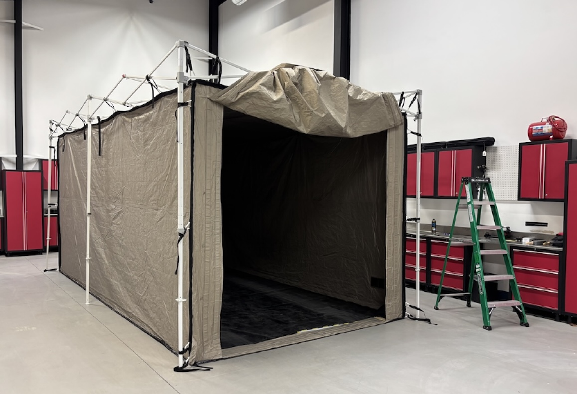 A large tan fabric Faraday tent on a white metal frame, its front flap rolled up to show a dark empty interior, set up inside a garage with red tool cabinets