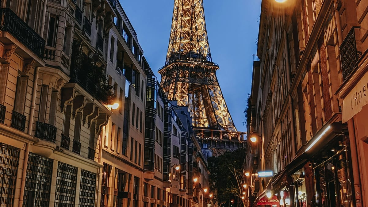 The Eiffel Tower lit up at dusk, seen down a Paris street lined with lit shopfronts